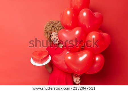 Horizontal shot of positive curly haired woman hides behind bunch of helium balloons holds heart cake comes to congratulate friend with birthday poses against vivid red background. Happy holiday