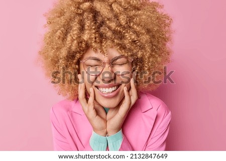 Headshot of happy young woman with curly hair keeps eyes closed hands on cheeks grins at camera wears transparent eyeglasses and formal jacket poses against pink background. Emotions concept