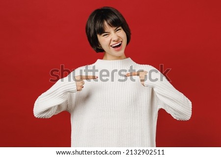Young surprised shocked amazed cool happy fun woman 20s wearing white knitted sweater point index finger on herself isolated on plain red color background studio portrait. People lifestyle concept.