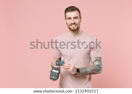 Smiling young bearded tattooed man guy in pastel t-shirt isolated on pink background. People lifestyle concept. Hold wireless modern bank payment terminal to process and acquire credit card payments