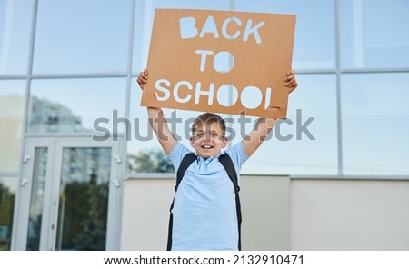 Positive schoolkid with backpack and poster with inscription Back To School standing against school building and looking at camera