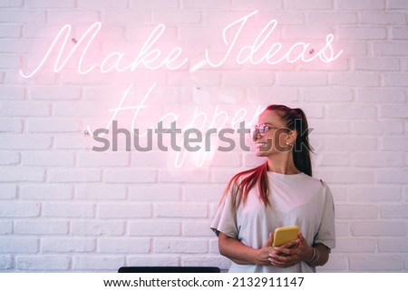 Happy Caucaisan hipster girl in stylish spectacles for provide eyes correction holding cellphone technology sitting under LED sign with winged motivation phrase - illuminated neon light as decoration