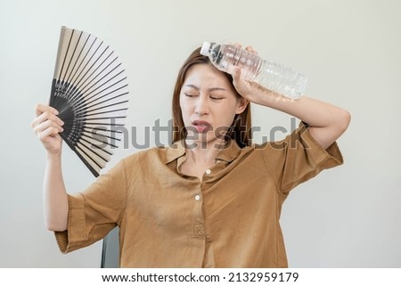 Summer heat stroke, hot weather, tired asian young woman sweaty and thirsty, refreshing with hand in blowing, wave fan to ventilation, holding cold water bottle tap her body when temperature high. Royalty-Free Stock Photo #2132959179
