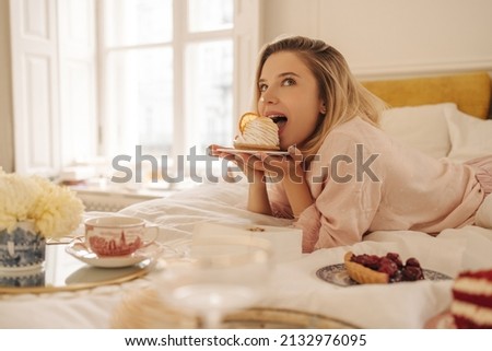 Lovely young caucasian girl appetizingly bites cake, looking up, lying in bed on her stomach. Beauty blonde woman in light pink pajamas is enjoying dessert. Sweets lover concept Royalty-Free Stock Photo #2132976095
