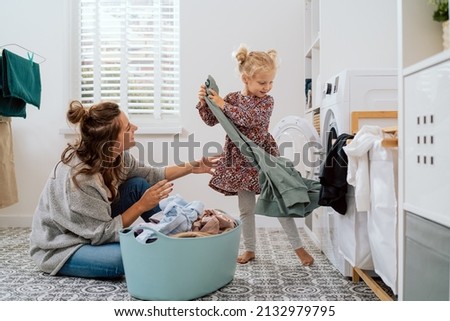 Mom spends time with daughter in bathroom, laundry room while doing daily chores, girl takes things out of washing machine and hands to woman who puts colorful clean clothes into bowl Royalty-Free Stock Photo #2132979795