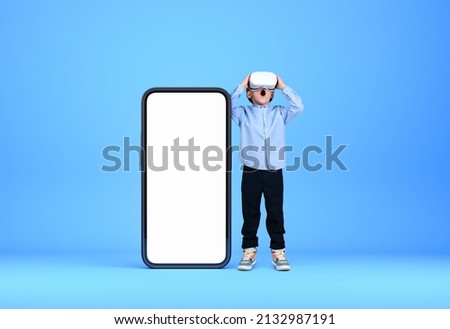 A little kid in virtual reality glasses, full length. Smartphone mock up copy space screen, blue background. Concept of metaverse and new device