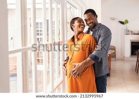 Lovely mid black man hugging his pregnant wife from behind standing near window at home. Happy middle aged black husband embracing pregnant woman while waiting for baby. African american mature couple Royalty-Free Stock Photo #2132995967