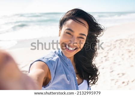 Portrait of smiling young woman taking a selfie at beach during. Cheerful hispanic woman enjoying at beach during holiday. Happy girl taking photo over exotic tropical beach looking at camera.