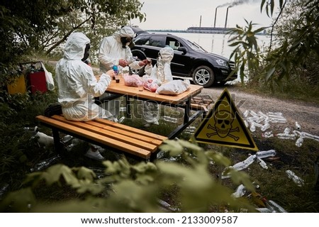 Environmentalists in radiation suits drinking in roadside forest with plastic bottles and poison toxic sign. Family resting at polluted territory with garbage and skull-and-crossbones warning symbol.