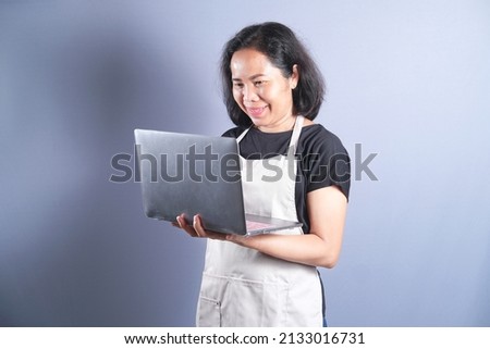 Asian housewife woman chef looks happy wearing apron holding laptop isolated over grey background. Copy space.