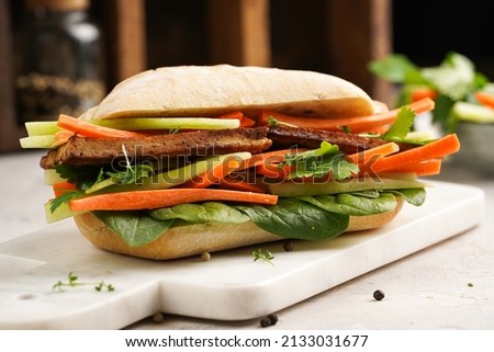 Traditional vietnamese banh mi sandwich with sliced smoked tofu, fresh spinach, shredded carrots and peeled cucumbers, cilantro on marble board on light grey textured background Royalty-Free Stock Photo #2133031677