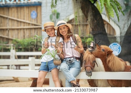 Asian mother and daughter feeding pony horse at animal farm. Outdoor fun for kids. Child feeds animal at pet zoo. Dwarf horses on the farm. Happy family petting a pony through wooden fence. Royalty-Free Stock Photo #2133073987