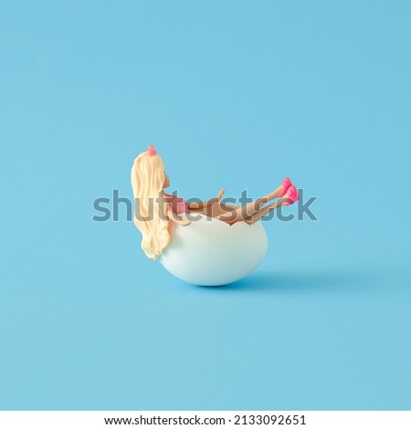 Blond girl lying in white egg shell against pastel blue background. Creative Easter concept. Minimal trendy composition.