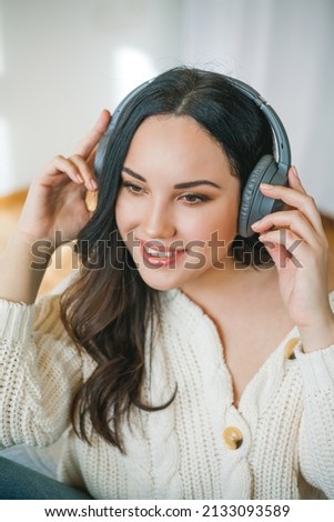Young beautiful dark-haired curly woman in a knitted cardigan listens to music on headphones while sitting in an armchair in a cozy home.