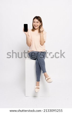 Happy Asian woman sitting and showing mobile phone and mini heart sign isolated over white background Royalty-Free Stock Photo #2133095255