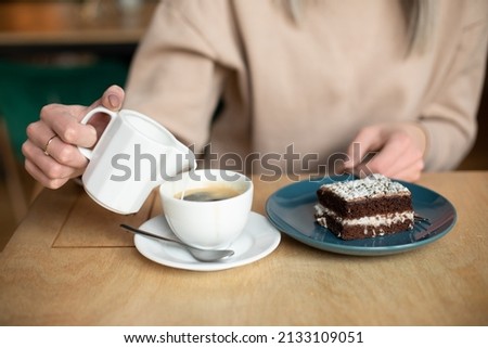 Cropped photo of blurred woman hands with ring, pouring milk cream in coffee cappuccino, latte. Coffee shop restaurant, sweet delicious chocolate cake brownie plate. Calm tranquil atmosphere, close up