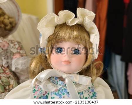 A closeup of the face of a doll. The doll has blue eyes, blond hair, and is wearing a white bonnet with a white ribbon around its face. Royalty-Free Stock Photo #2133130437