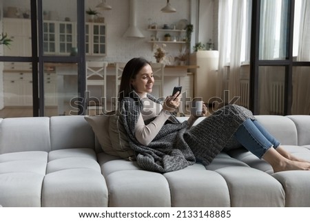 Peaceful young female relaxing on cozy sofa covered with knitted warm plaid, holding smart phone and teacup drinking tea spend carefree free time alone at home. Weekend leisure, modern tech concept Royalty-Free Stock Photo #2133148885