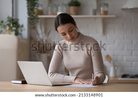 Young woman sit at table with laptop in kitchen holds pen, take notes on paper, writes letter. Student make research, prepare for exam at home. Jotting information, create to-do list, planning concept Royalty-Free Stock Photo #2133148901