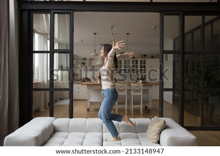 Young woman have fun alone at home, celebrate relocation day to own or rented modern apartment, raise arms dance, jumping barefoot on sofa in living room feel happy, looks untroubled. Tenancy concept Royalty-Free Stock Photo #2133148947