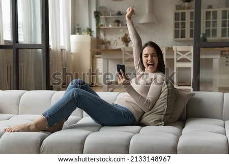 Woman hold cellphone sit on sofa, express excitement read unbelievable great news get profitable offer, enjoy moment of victory raise hand scream with joy feel happy. Success, lottery, bet win concept Royalty-Free Stock Photo #2133148967