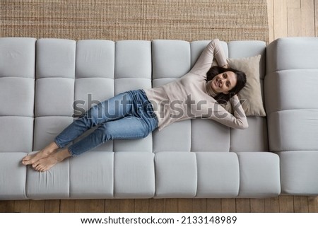Overhead view serene young woman put hands behind head relaxing on fashionable soft sofa, smile look at camera. Carefree leisure on weekend at modern home, fresh conditioned air, comfort life concept Royalty-Free Stock Photo #2133148989