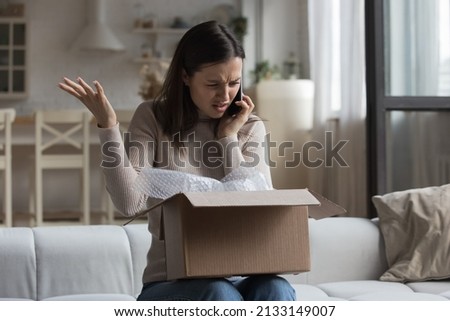 Woman sit on sofa open parcel box looks inside check purchased damaged items feels angry, complaining speaks on smartphone to customer support. Broken goods, bad services, dissatisfied client concept Royalty-Free Stock Photo #2133149007