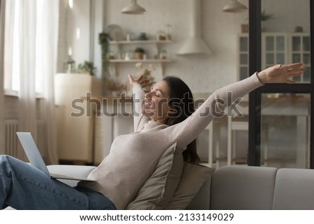 Happy young woman relaxing alone on cozy sofa put wireless computer on laps, raising her arms, female smiling with eyes closed enjoy carefree untroubled weekend, accomplish online task feeling free Royalty-Free Stock Photo #2133149029