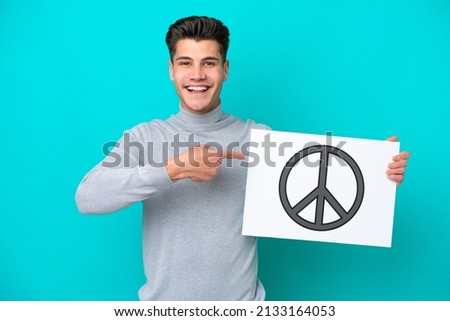 Young handsome caucasian man isolated on blue bakcground holding a placard with peace symbol and  pointing it
