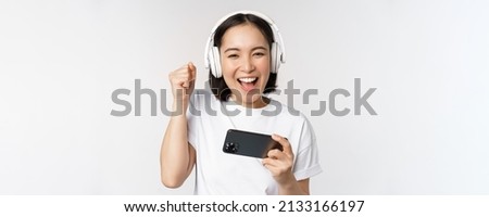 Happy asian girl gamer, playing on mobile phone, watching on smartphone, wearing headphones, standing over white background