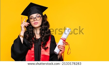 Girl graduate in graduation hat and eyewear with diploma on yellow background. Self-confident young woman wearing graduation cap and ceremony robe holding Certificate tied with red ribbon. Education Royalty-Free Stock Photo #2133178283