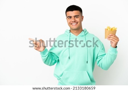 Young handsome man holding fried chips over isolated white background pointing finger to the side