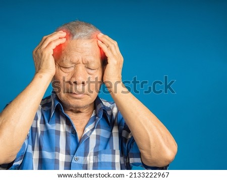 Senior man suffering headache pain while standing against a blue background. Aged people and healthcare concept