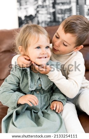 Portrait of adorable little brother and sister sitting on chair, hugging and smiling. Cute stylish dressed small kids having fun, playing together and laughing, childhood concept. High quality photo