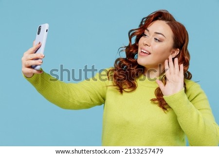 Young smiling happy chubby overweight plus size big fat fit woman wearing green sweater doing selfie shot on mobile cell phone waving hand isolated on plain blue background. People lifestyle concept