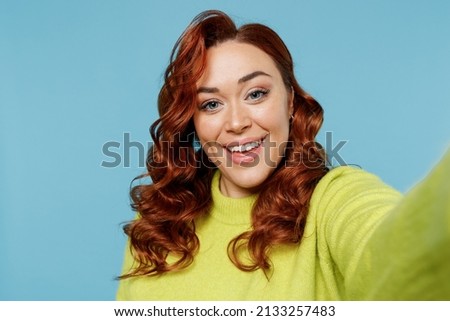 Young close up smiling chubby overweight plus size big fat fit woman wear green sweater do selfie shot pov on mobile phone isolated on plain blue background studio portrait. People lifestyle concept