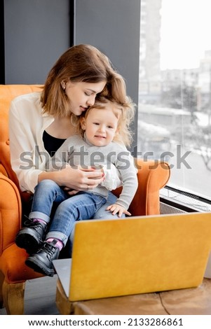 Young beautiful mother sitting at chair with little lovely daughter, having video call from family, smiling. Attractive woman with cute toddler using laptop, watching cartoons, concept of childhood.