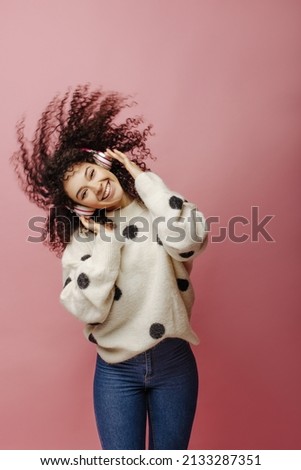 Happy young caucasian woman in headphones waving her hair on pink background. Brunette girl with curly hair listens to music. Audio technology, concept