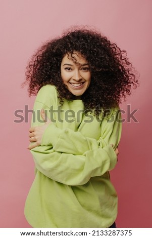 Cute curly young caucasian woman happily hugging herself while looking at camera on pink background. Brunette in light green hoodie feels comfort. Lifestyle, different emotions, leisure concept.