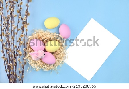 Nest with multi-colored eggs and an Easter bunny on a blue background next to willow branches and a white sheet for text. Easter picture with copy space.