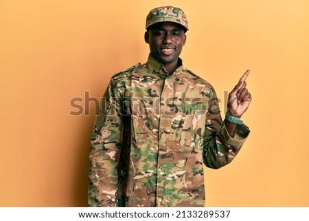 Young african american man wearing army uniform with a big smile on face, pointing with hand finger to the side looking at the camera. 