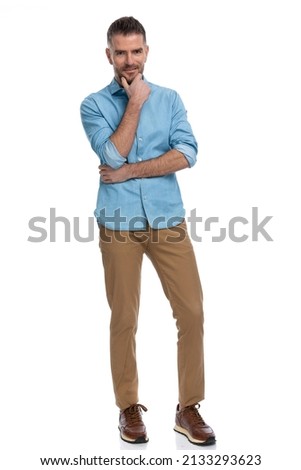 full body picture of young casual man in denim shirt crossing arms, holding hand to chin and thinking, being creative on white background in studio