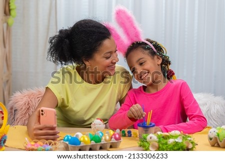 Mom and daughter with funny bunny ears talk on a video call using the phone. African American woman and little girl are sitting at table in festively decorated room. Happy easter. Close up.