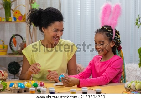 Mom and daughter with funny bunny ears gluing pictures on colored eggs. African American woman and little girl are sitting at table in festively decorated room at home. Happy easter. Close up.