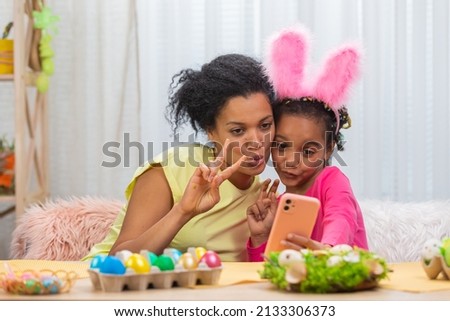 Mom and daughter with funny bunny ears take selfies and have fun. African American woman and little girl are sitting at table in festively decorated room at home. Happy easter. Close up.