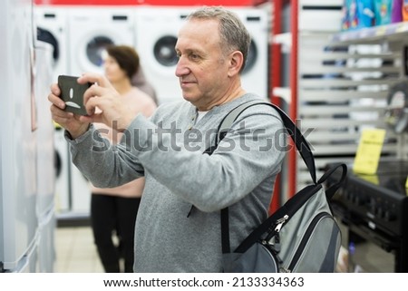 Mature man standing in salesroom of appliance store and photographing refrigerator with his smartphone.