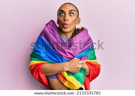 Hispanic man wearing make up and long hair wrapped in rainbow lgbtq flag making fish face with mouth and squinting eyes, crazy and comical. 
