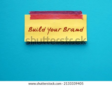 Yellow note stick on blue copy space background with handwritten text BUILD YOUR BRAND, means entrepreneurs differentiating service or product  from competition by creating brand that attract customer
