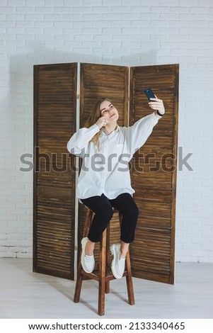 Emotional young woman in a white cotton shirt gesticulates and looks at the screen of a modern gadget. A girl with a mobile phone takes a selfie