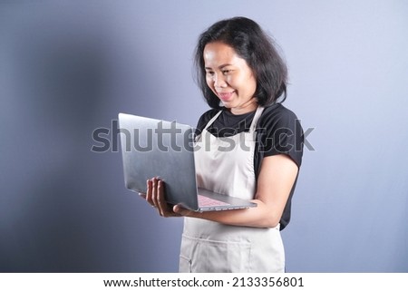 Asian housewife woman chef looks happy wearing apron holding laptop isolated over grey background. Copy space.